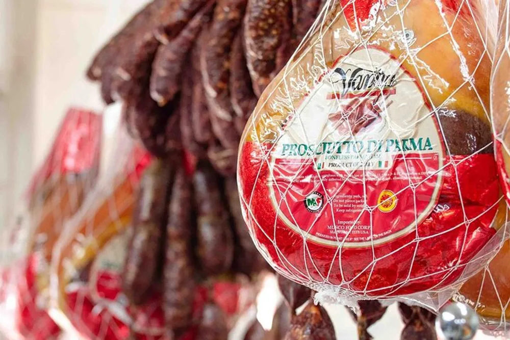 The image shows a selection of cured meats prominently featuring a Prosciutto di Parma hanging and packaged for sale