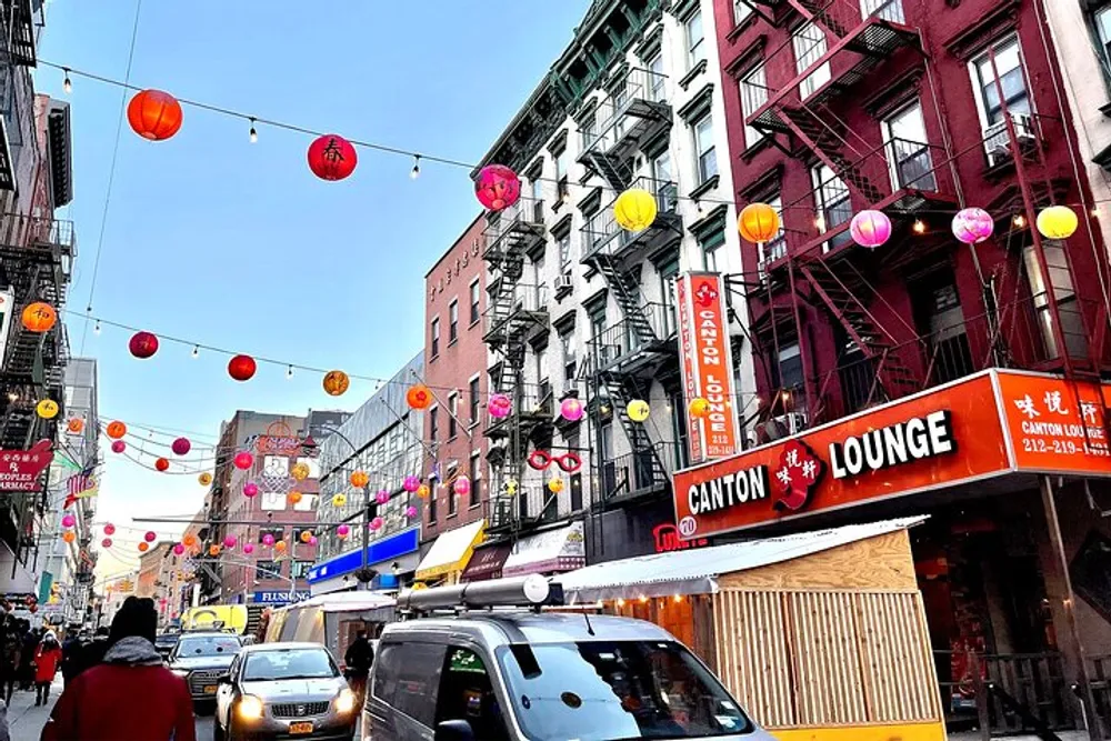 Colorful lanterns are strung across a lively urban street lined with businesses including a place called Canton Lounge bustling with pedestrian and vehicular activity under a clear sky
