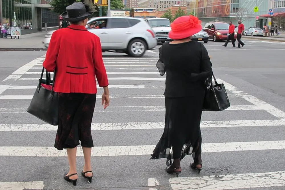 Two elegantly dressed individuals in red and black attire with matching hats are waiting to cross a bustling city street