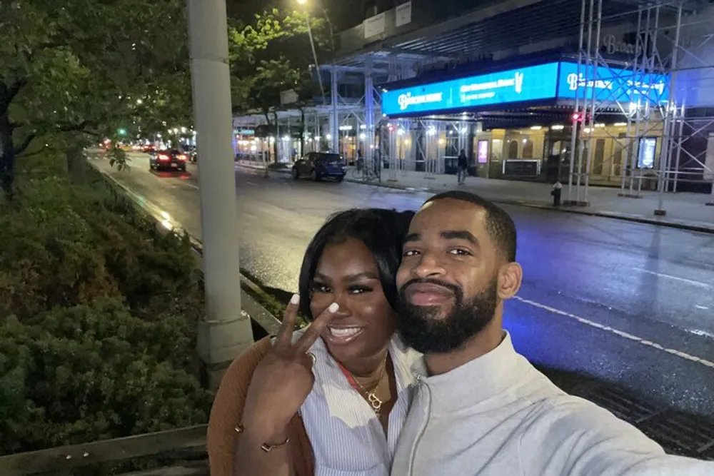 A happy couple is taking a selfie on a city street at night with the woman showing a peace sign