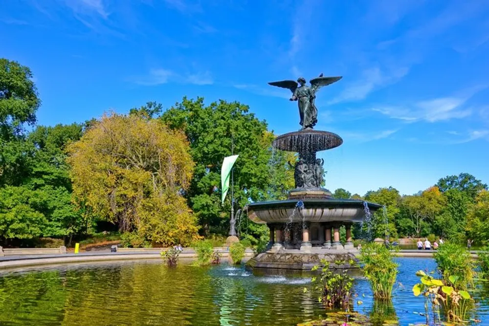 The image shows the Bethesda Fountain located in Central Park New York City featuring the Angel of the Waters statue atop with a clear blue sky in the background and lush green trees surrounding the area