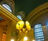 The image features the iconic golden clock in the center of the main concourse of Grand Central Terminal with its opulent architectural details and soft ambient lighting
