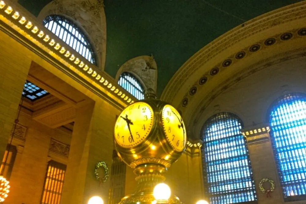The image features the iconic golden clock in the center of the main concourse of Grand Central Terminal with its opulent architectural details and soft ambient lighting