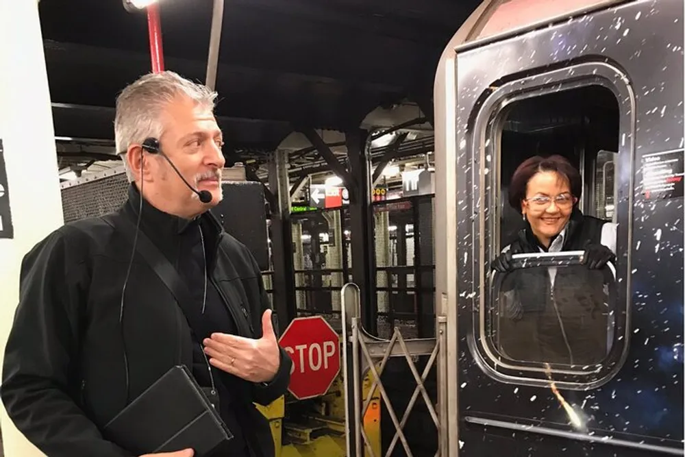 A man is standing at a subway platform next to a stopped train communicating with the female train operator through the window
