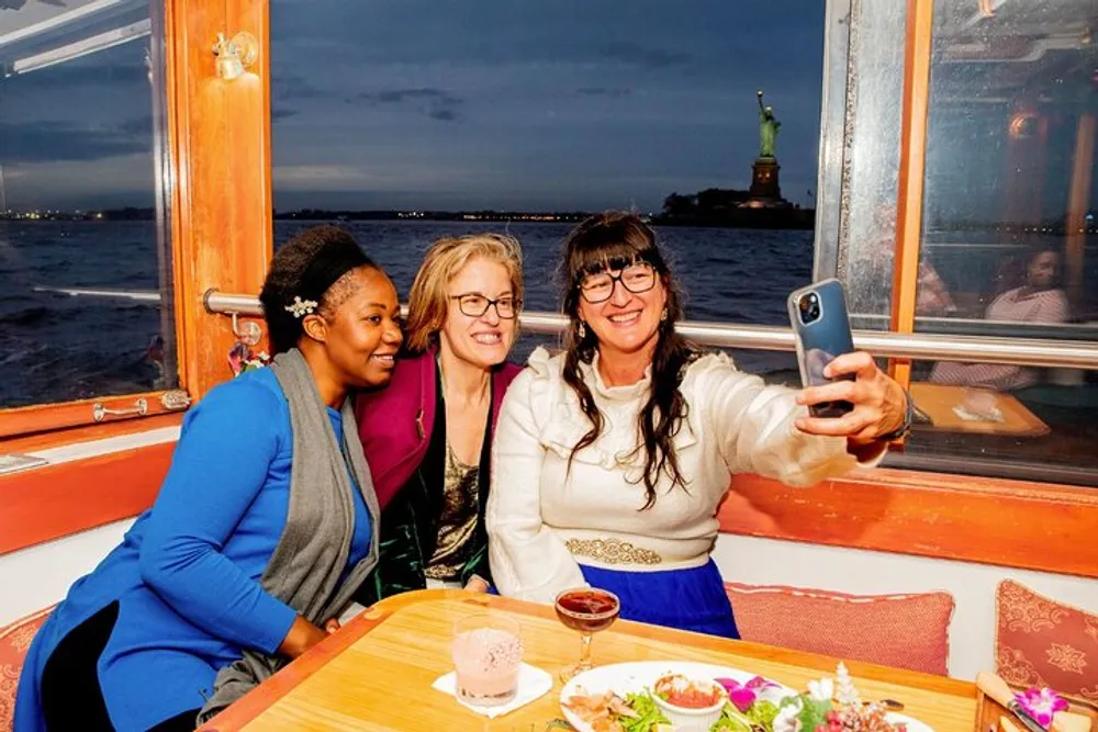 Three people are taking a selfie on a boat with the Statue of Liberty in the background at dusk