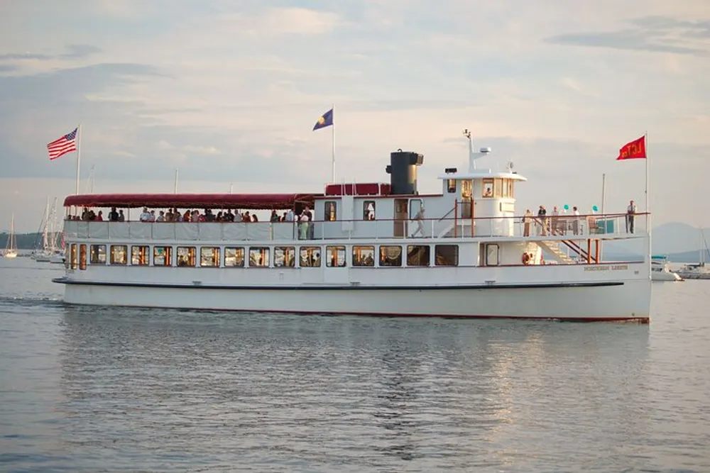 The image shows a two-deck riverboat with passengers on both levels cruising on calm waters with other boats and the soft glow of the evening sky in the background