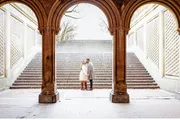 A couple shares an affectionate moment under an ornate arched passageway with a staircase in the background.