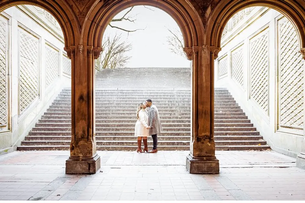 A couple shares an affectionate moment under an ornate arched passageway with a staircase in the background