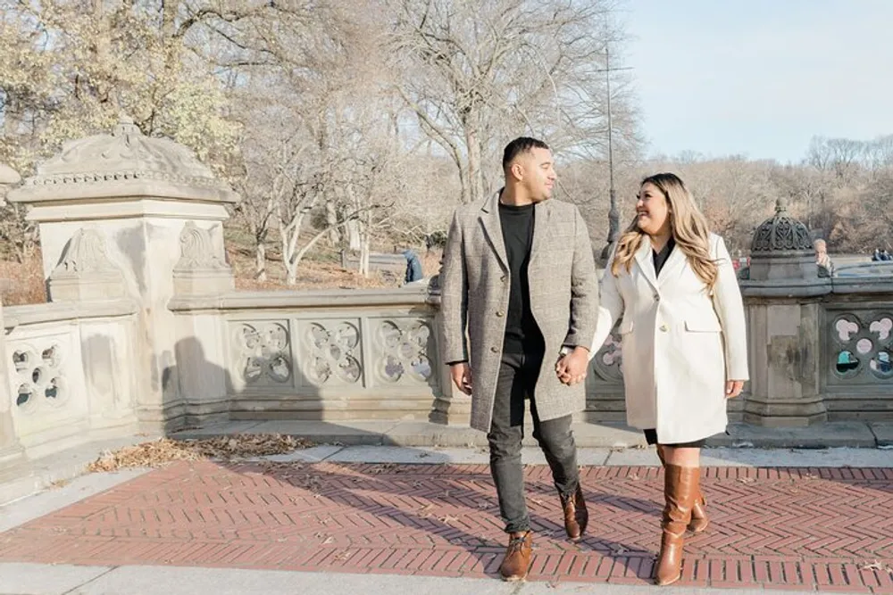 A smiling couple is holding hands and looking at each other while walking along a brick path near an ornate stone balustrade in a park setting with bare trees in the background