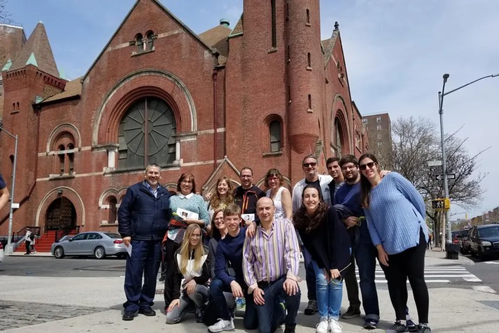 A group of smiling people poses together in front of a large red brick church on a sunny day with light cloud cover