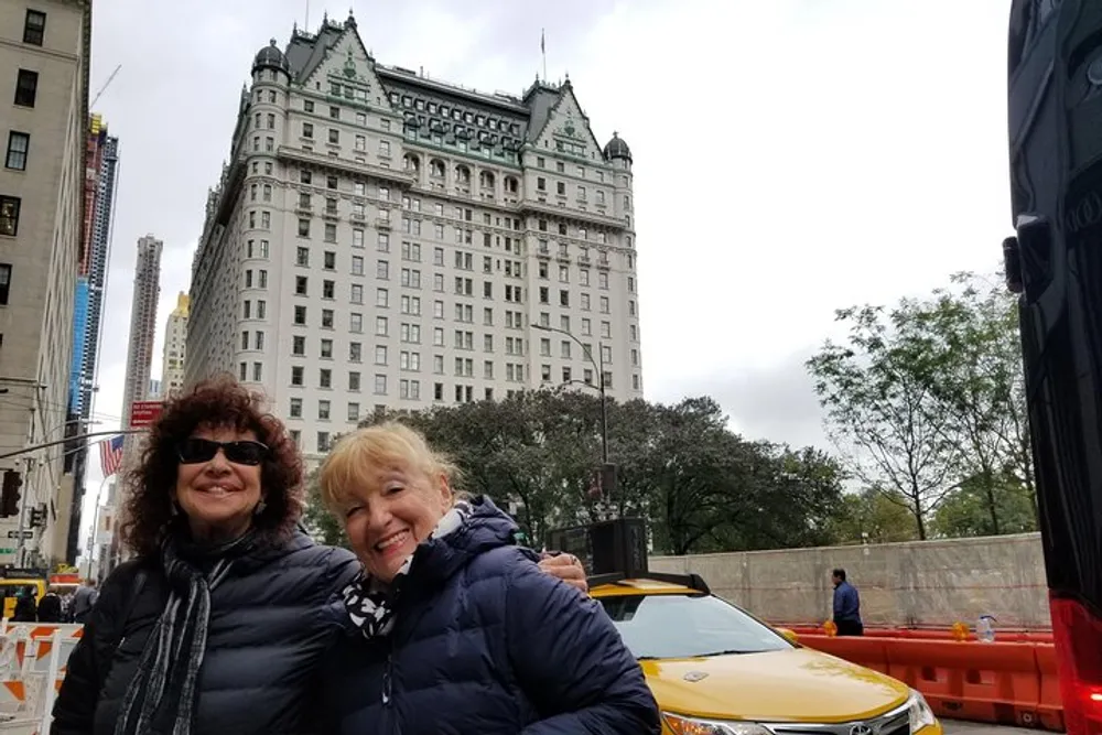 Two smiling women pose in front of an impressive historic-looking building with distinctive architectural features with a yellow taxi and construction barriers also visible in the scene