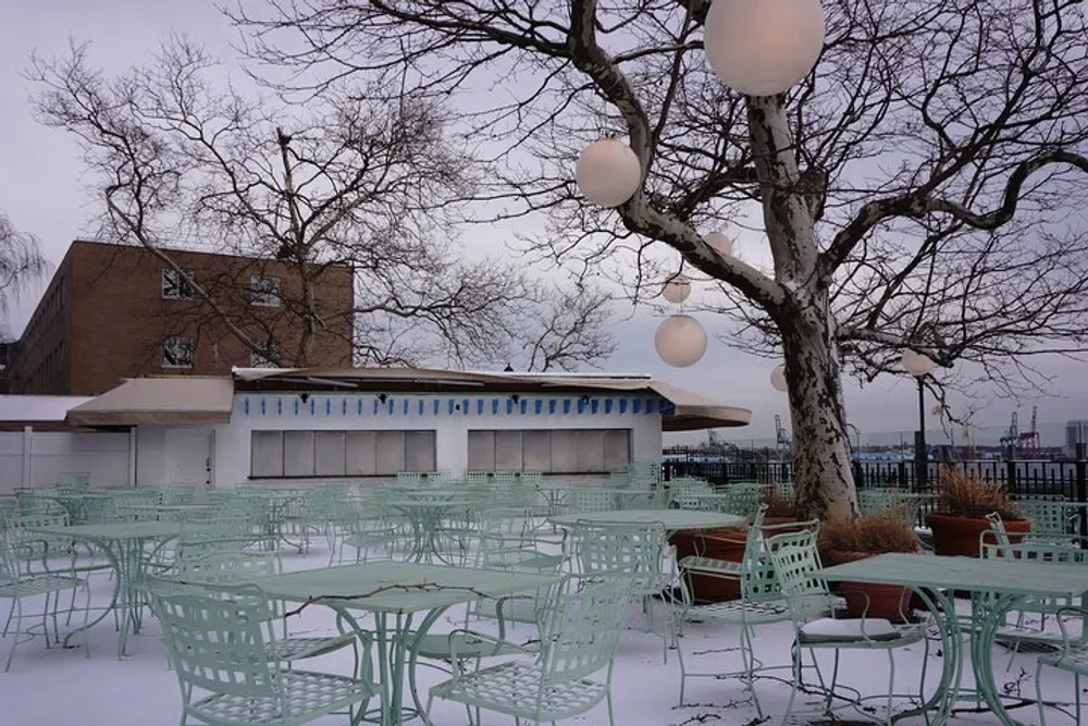 An outdoor seating area with green metal chairs and tables is covered in snow framed by a leafless tree and spherical light fixtures with a building and overcast sky in the background