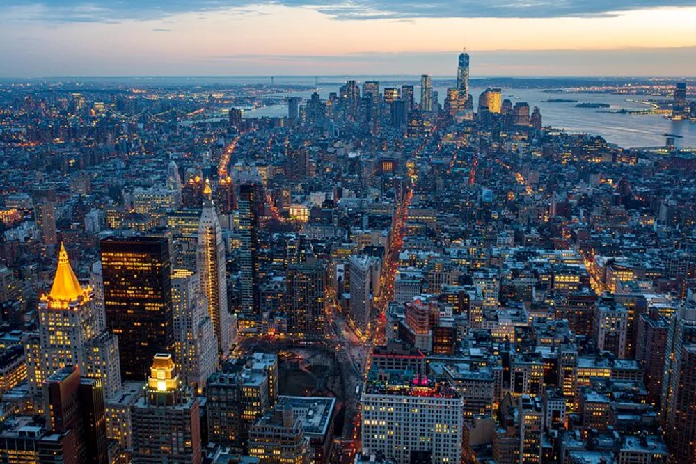 An aerial view of New York City during twilight showcases the densely packed buildings and streets with lights starting to illuminate the bustling urban landscape