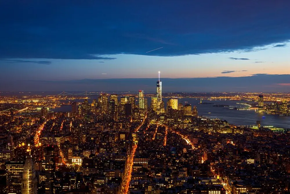The image captures an aerial view of a vibrant city skyline at twilight with lights illuminating the streets and buildings and a prominent skyscraper stands out against the fading blue sky