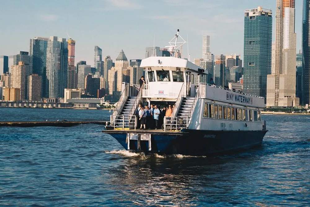 A ferry marked NY Waterway is on the water with passengers standing on the deck against the backdrop of a city skyline