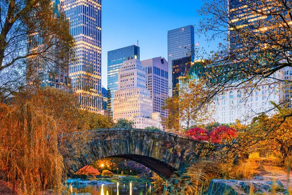 The image shows a picturesque view of a stone bridge in Central Park with the colorful autumn foliage in the foreground and the illuminated skyscrapers of New York City in the background at dusk