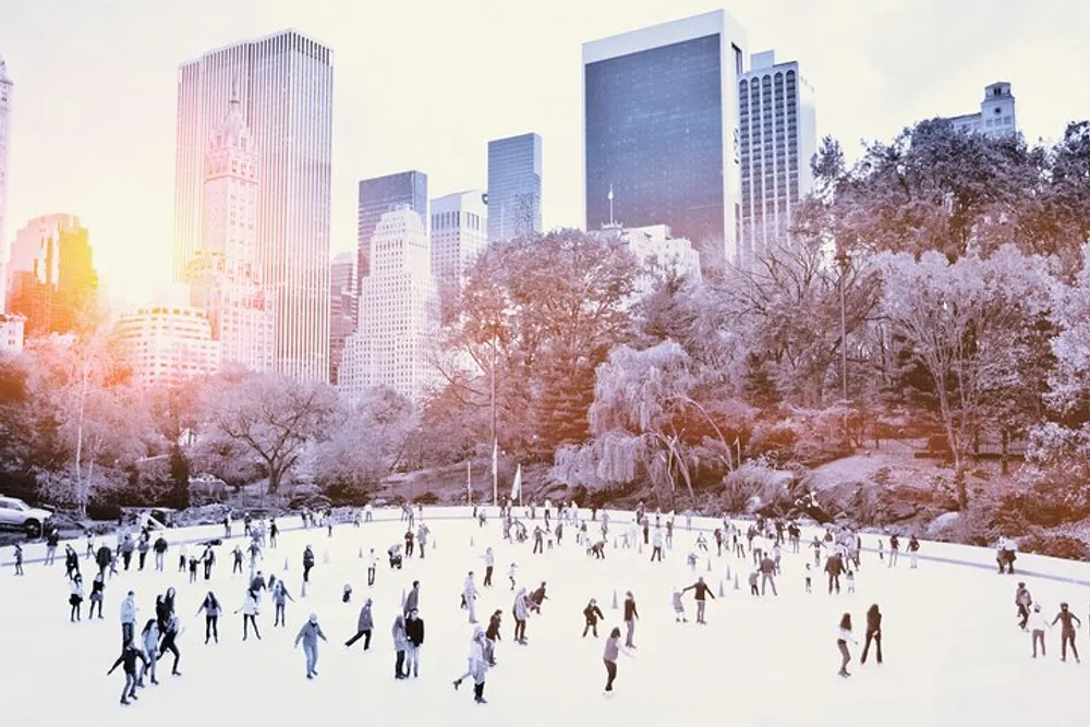 People are ice skating at a rink surrounded by trees with a backdrop of city skyscrapers bathed in a soft warm light