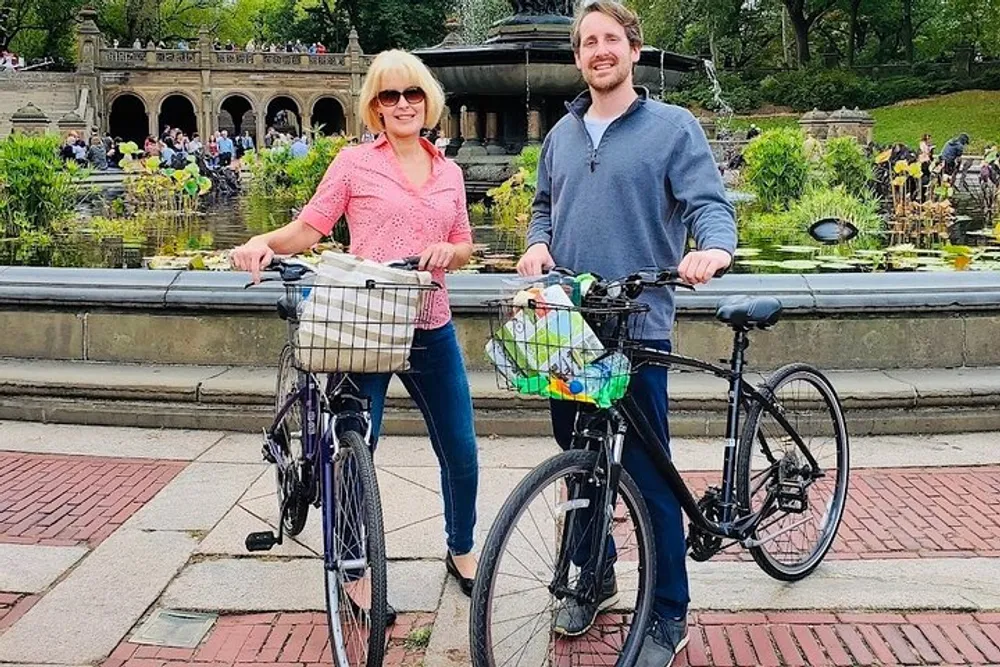 A man and a woman are smiling as they stand with their bicycles in front of a scenic pond and a decorative stone archway in a lush park setting