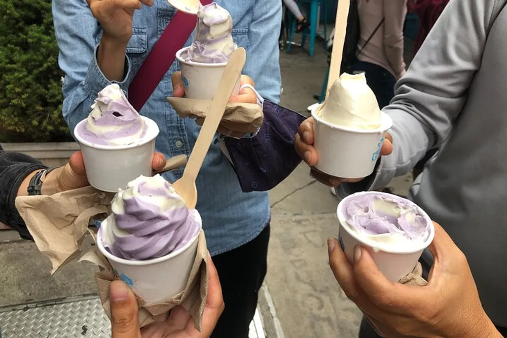 Several people are holding cups of swirled soft-serve ice cream predominantly in shades of purple and white with wooden spoons