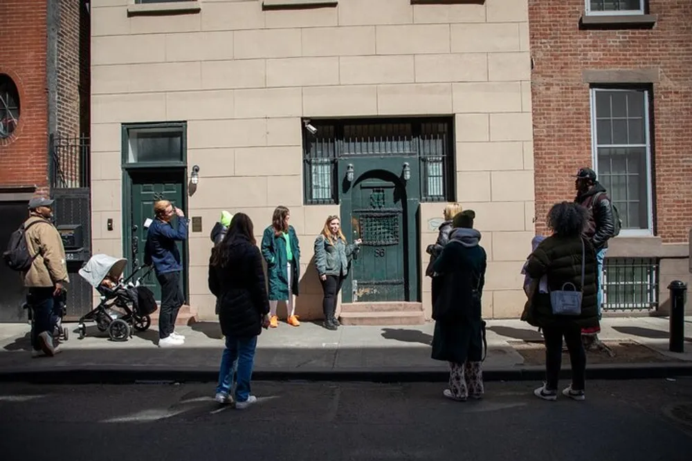 A group of people some standing on the sidewalk and others crossing the street gather in front of a building with a green door possibly on a guided tour or a casual group meeting