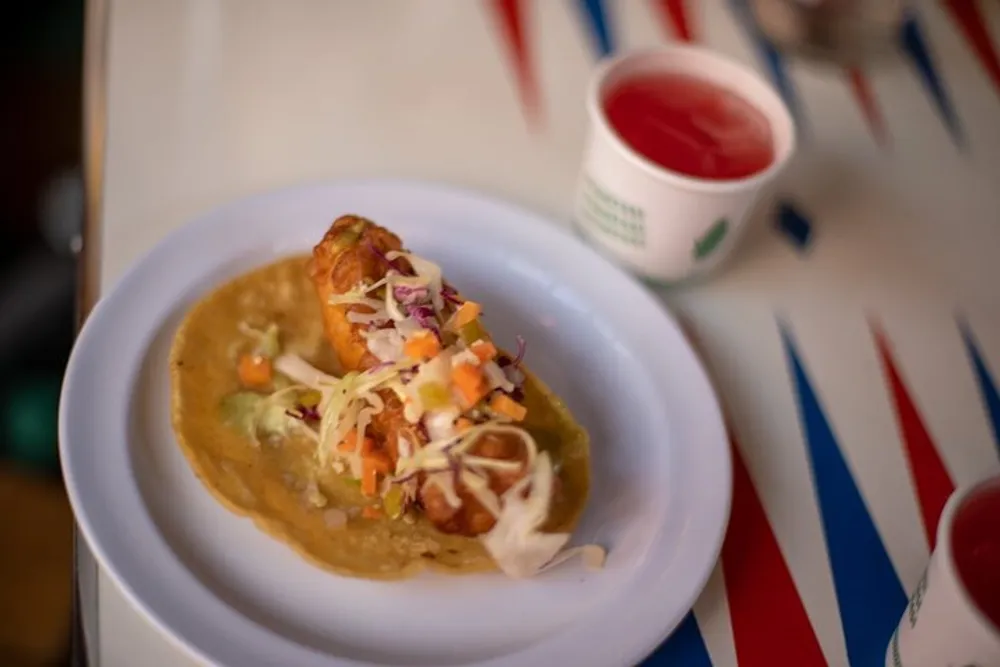 A taco with a crispy shell is served on a white plate with a red drink in the background which is placed on a table with a striped red white and blue tablecloth