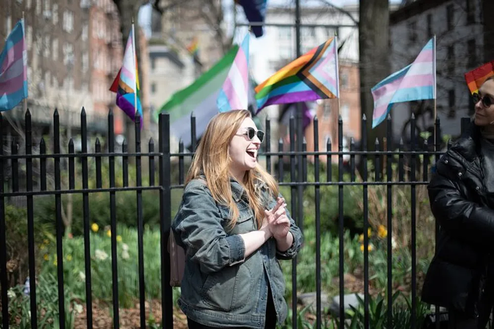 A smiling individual clapping hands with sunglasses on is standing in front of a fence with several pride flags fluttering in the background