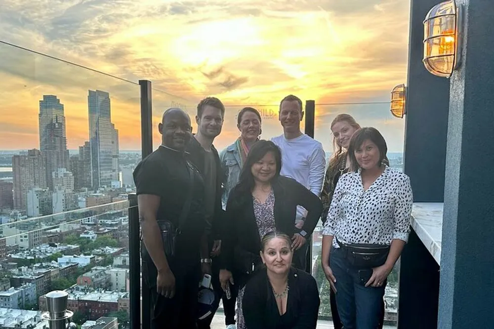 A group of eight people poses for a photo on a rooftop with a cityscape and sunset in the background