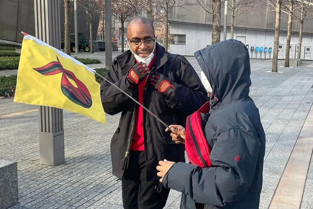 Two individuals one holding a flag with a red and yellow emblem are engaged in a conversation outdoors