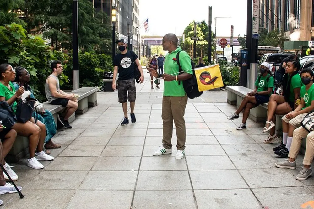 A person in a green shirt appears to be speaking to a group of attentive listeners outdoors near a city street
