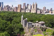 An aerial view of Belvedere Castle amidst the lush greenery of Central Park with the Manhattan skyline in the background.