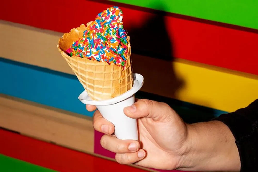 A person is holding a waffle cone with a vanilla ice cream topped with rainbow sprinkles against a colorful striped background