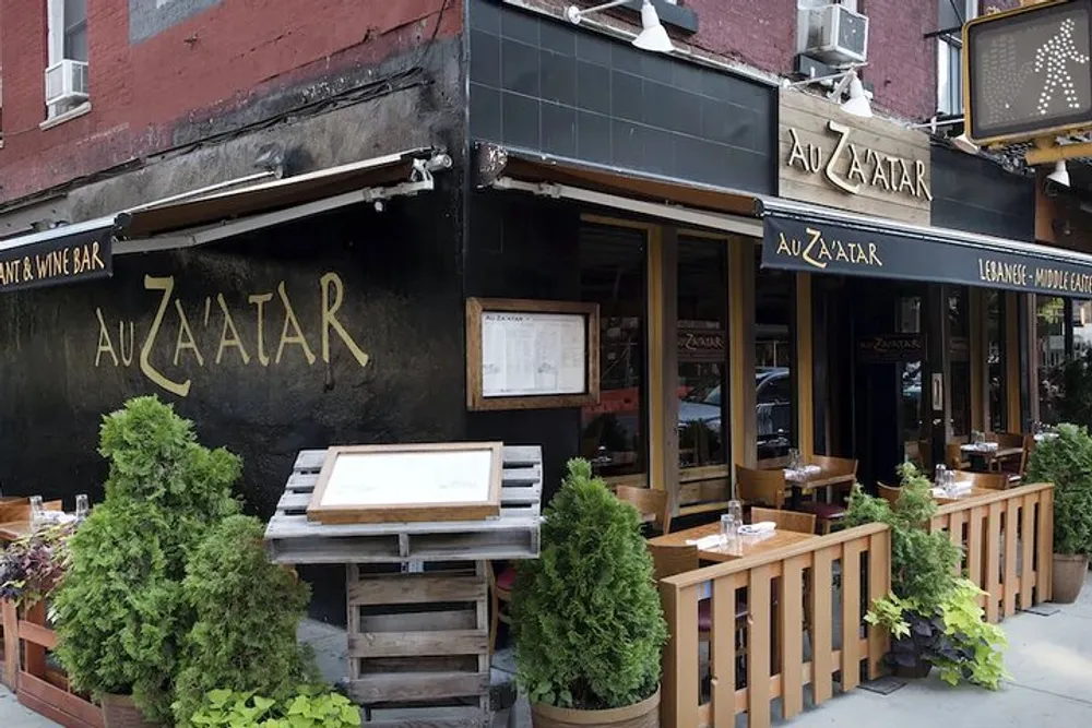 The image shows the exterior of Au Zaatar a LebaneseMiddle Eastern restaurant featuring sidewalk seating enclosed by wooden barriers and surrounded by potted green plants