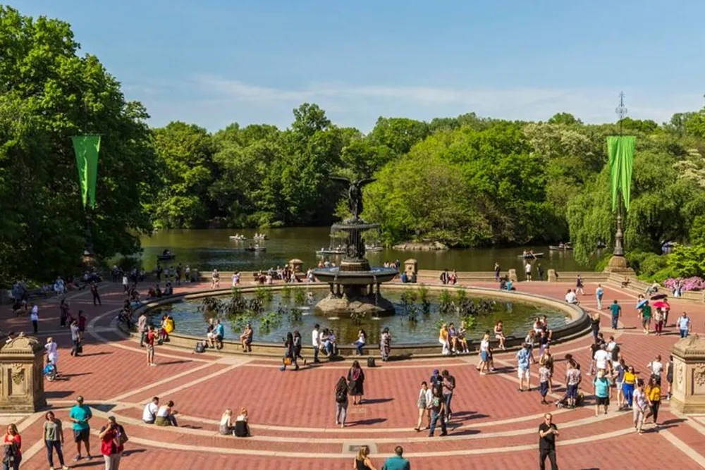 The image shows a bustling scene at a circular outdoor plaza with a large fountain in the center surrounded by people and greenery under a clear sky