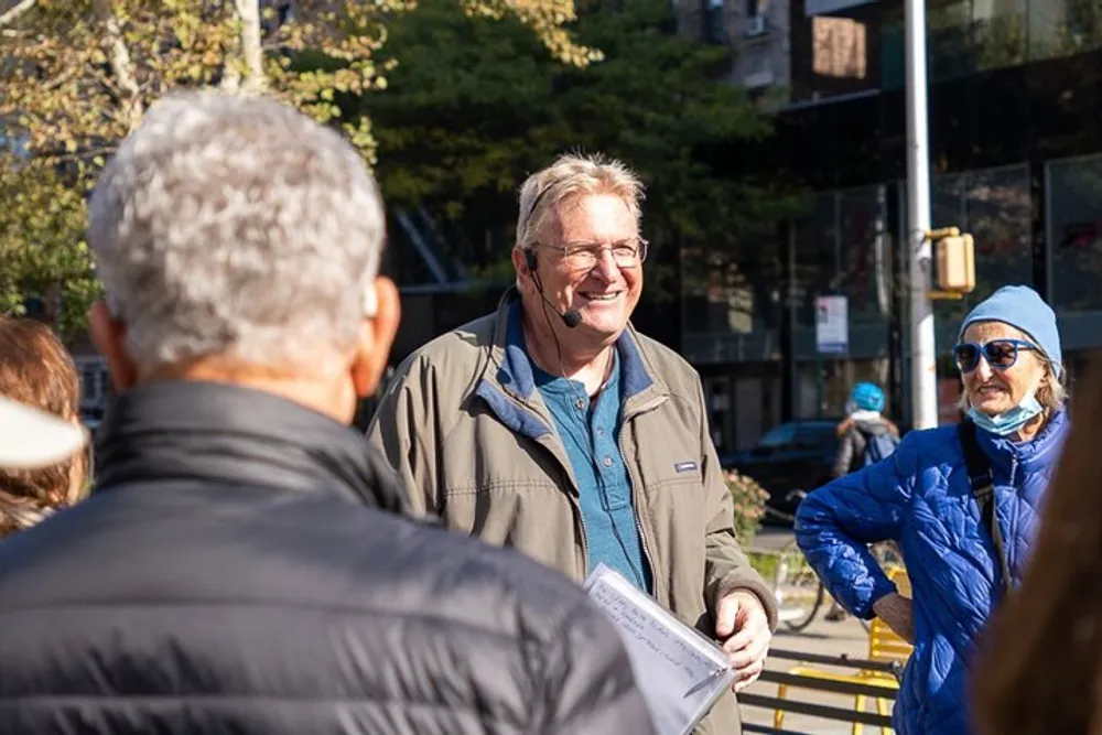 A man with glasses and a headset is smiling as he appears to lead or participate in an outdoor group discussion in a city setting