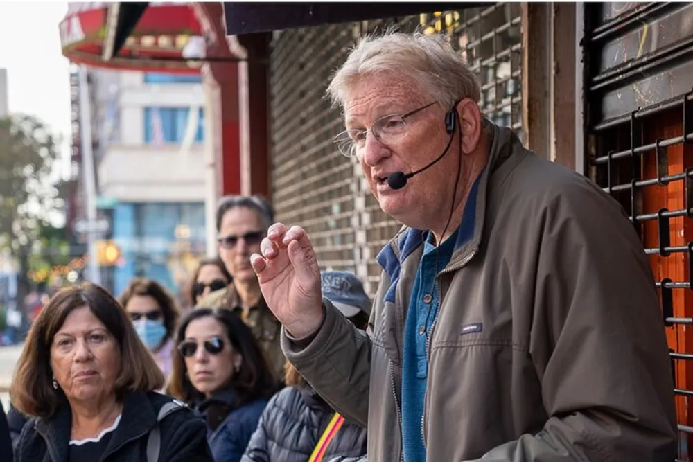 An older man wearing a headset microphone is speaking animatedly on a city street while a group of attentive people listen behind him