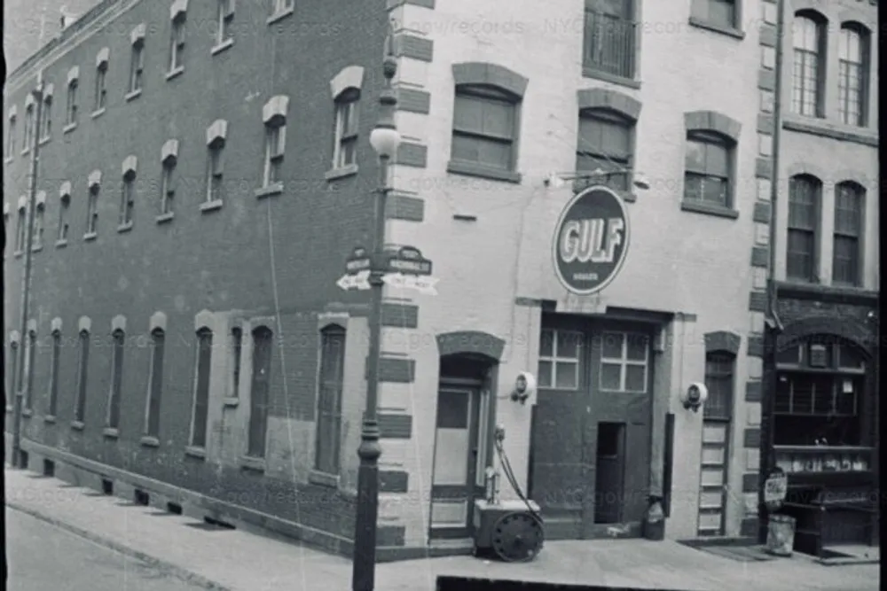 The image shows a vintage black and white photo of an old city street corner with a Gulf gas station sign brick buildings and a classic street lamp