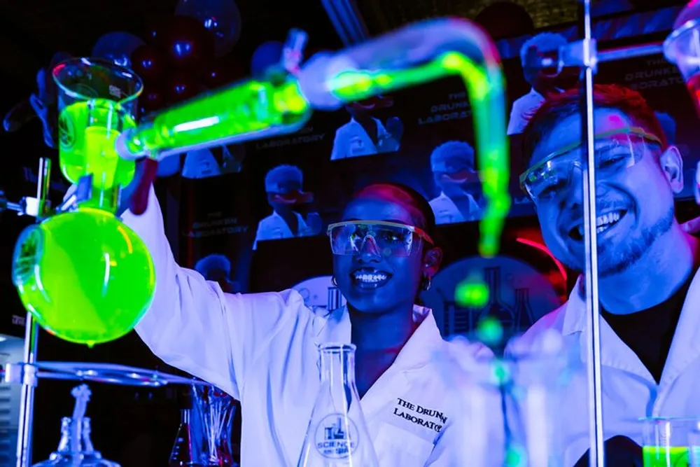 Two people wearing lab coats and safety glasses are working with glowing green chemicals in a laboratory setting