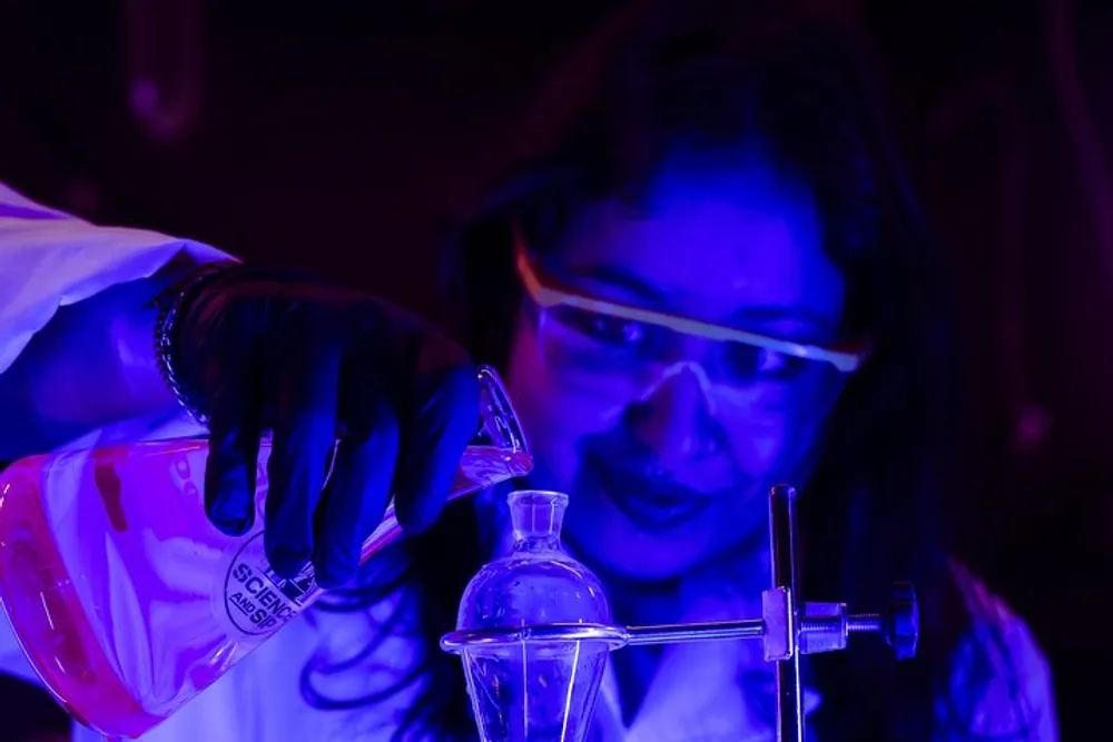 A person wearing safety goggles and gloves is conducting an experiment by pouring a substance from a beaker into a flask under blue lighting in a laboratory setting