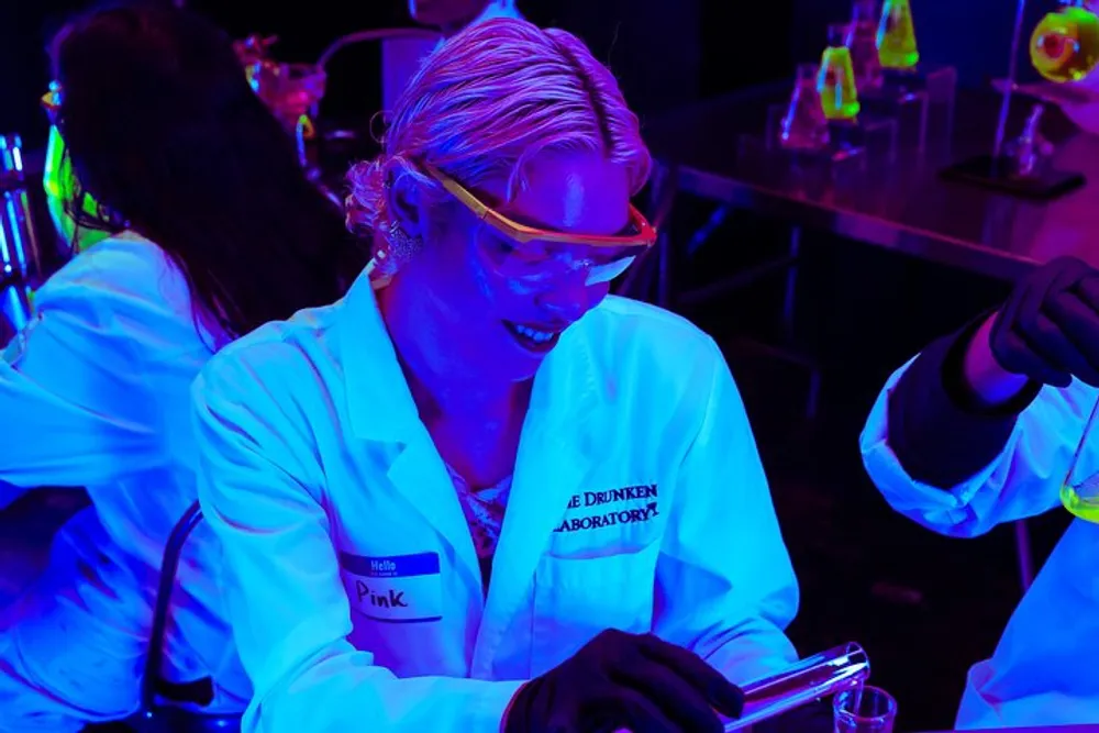 A person wearing protective goggles and a lab coat labeled The Drunken Laboratory is engaged in an experiment with colorful liquids under a blacklight