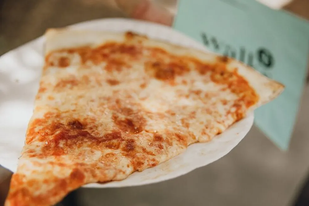 A person is holding a large slice of cheese pizza on a paper plate