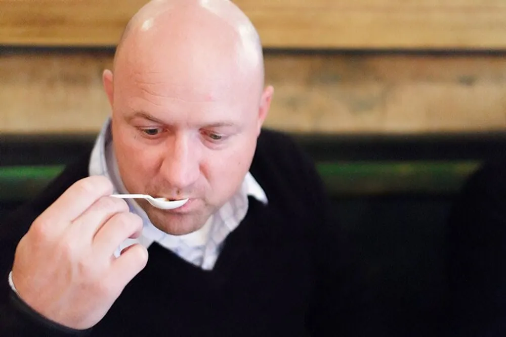A bald man in a black sweater is eating with a fork against a blurry wooden background