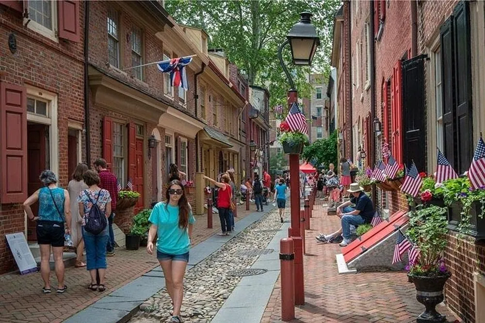 Visitors stroll down a quaint cobblestone street lined with brick buildings adorned with American flags indicative of a patriotic celebration or a historical district