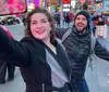 A woman takes a selfie with a joyful man photobombing in the background while they are both in a bustling urban setting with bright city lights