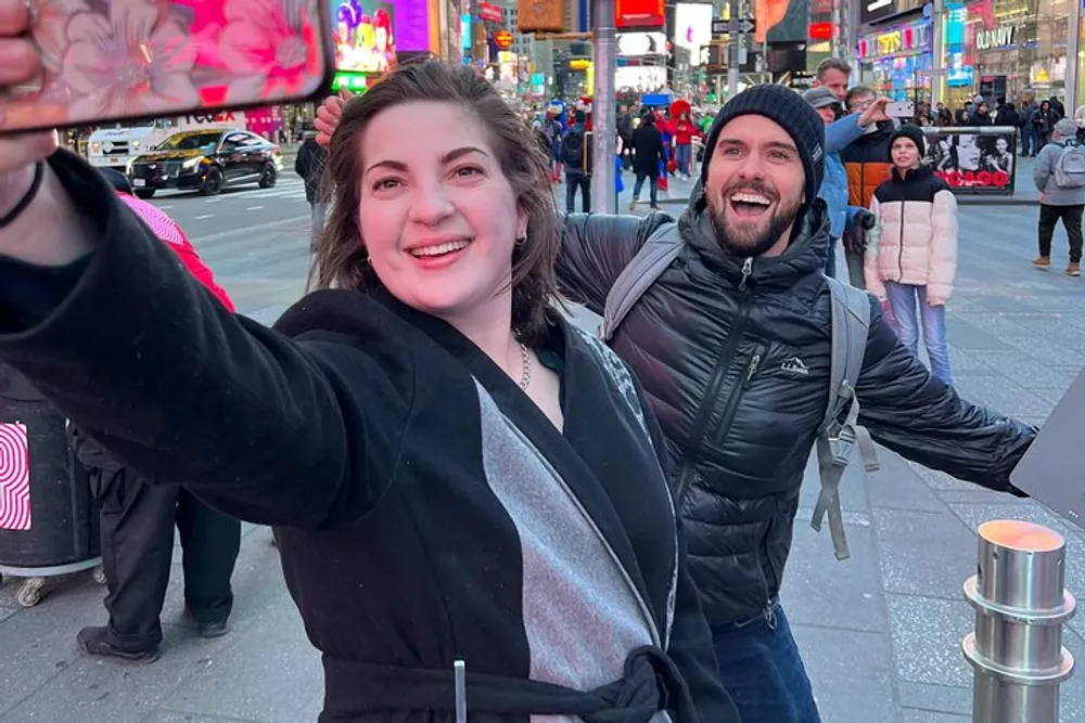 A woman takes a selfie with a joyful man photobombing in the background while they are both in a bustling urban setting with bright city lights