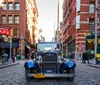 A vintage car is parked on a cobblestone street in an urban area with pedestrians and classic architecture creating a scene that blends historical and modern elements