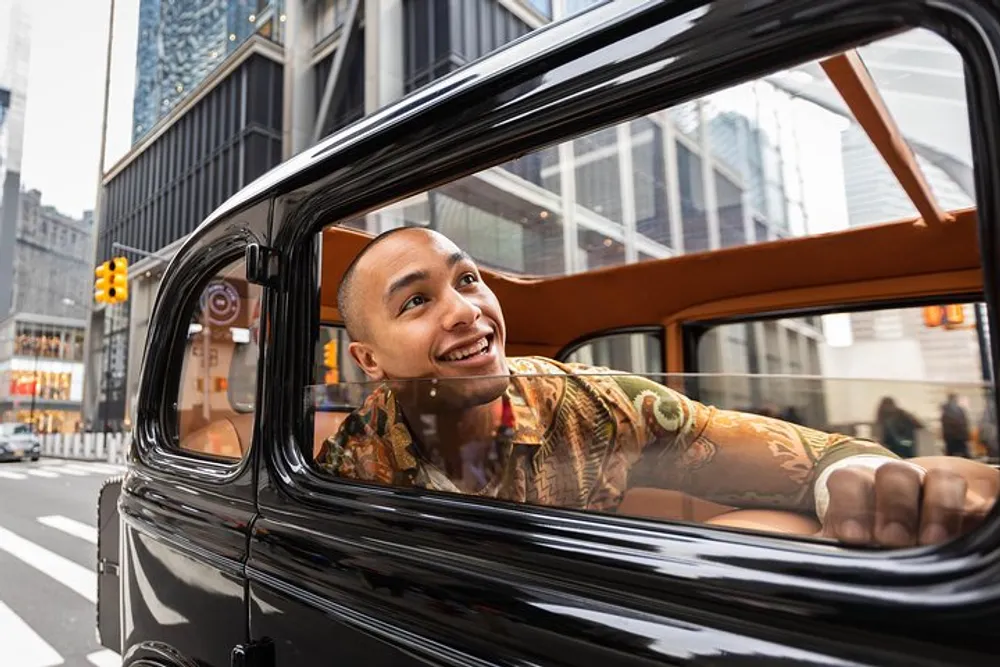 A person is peering out from the rear window of a classic black vehicle with a cheerful expression amidst an urban backdrop with high-rise buildings and street signs