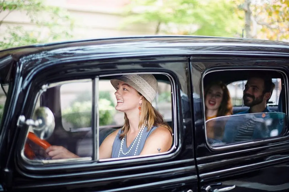 Three people are smiling and enjoying a ride in a vintage car with the driver wearing a hat and the passengers visibly happy in the back seat