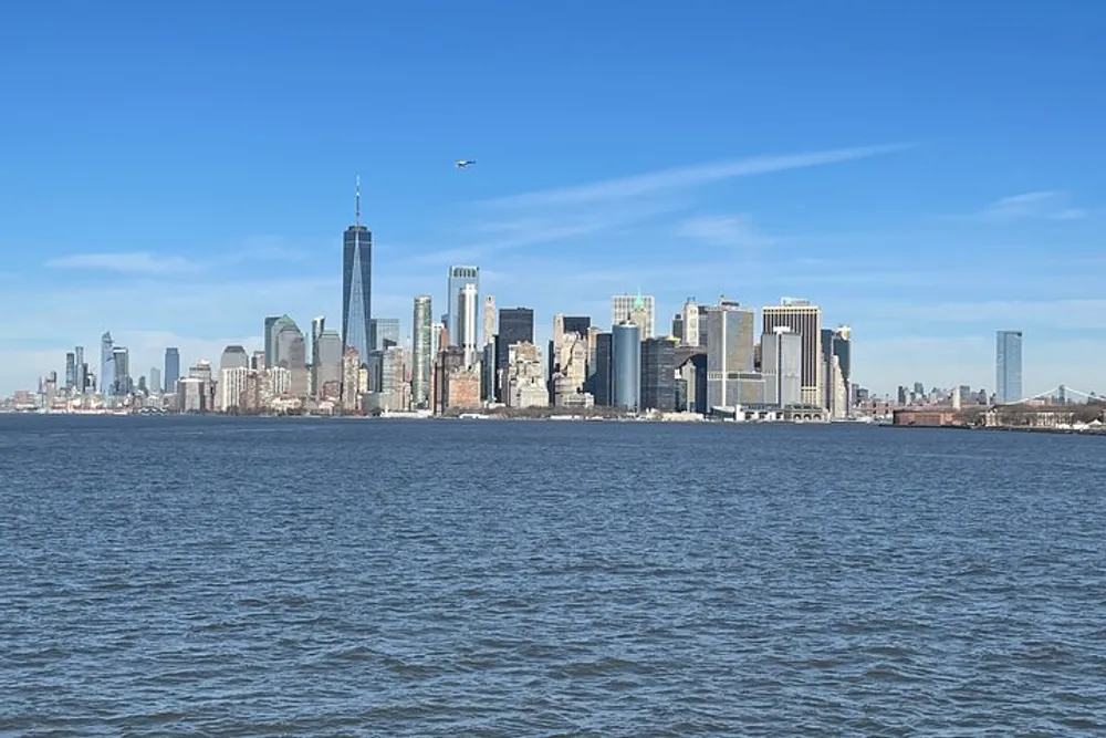 This image shows a clear daytime view of the Manhattan skyline with the One World Trade Center standing prominently as seen from across a body of water possibly the Hudson River under a mostly clear blue sky with a helicopter flying in the distance
