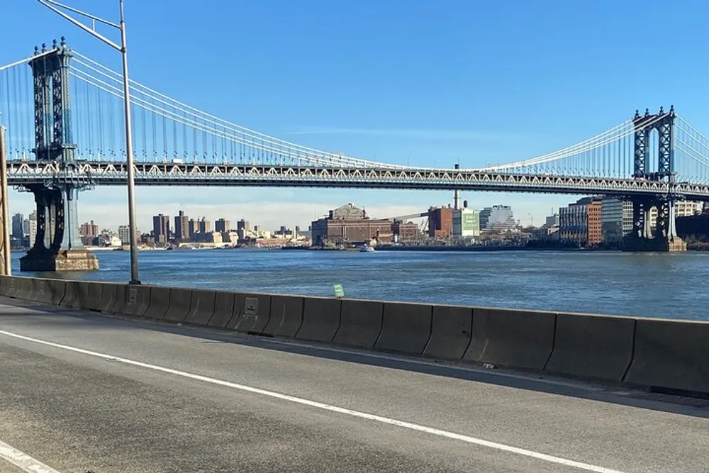 The image shows a sunny view of the Manhattan Bridge spanning over the East River with the New York City skyline in the background