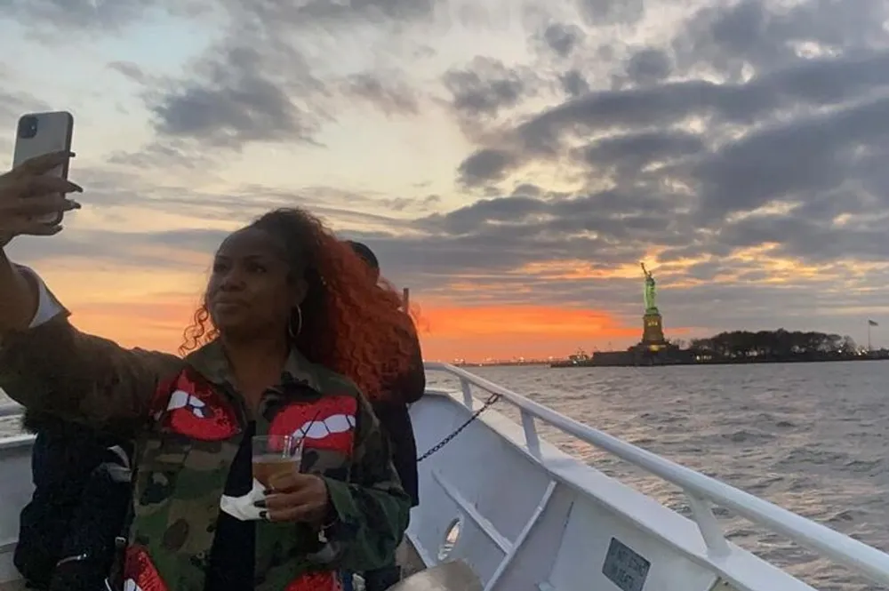 A person is taking a selfie on a boat with the Statue of Liberty in the background at sunset